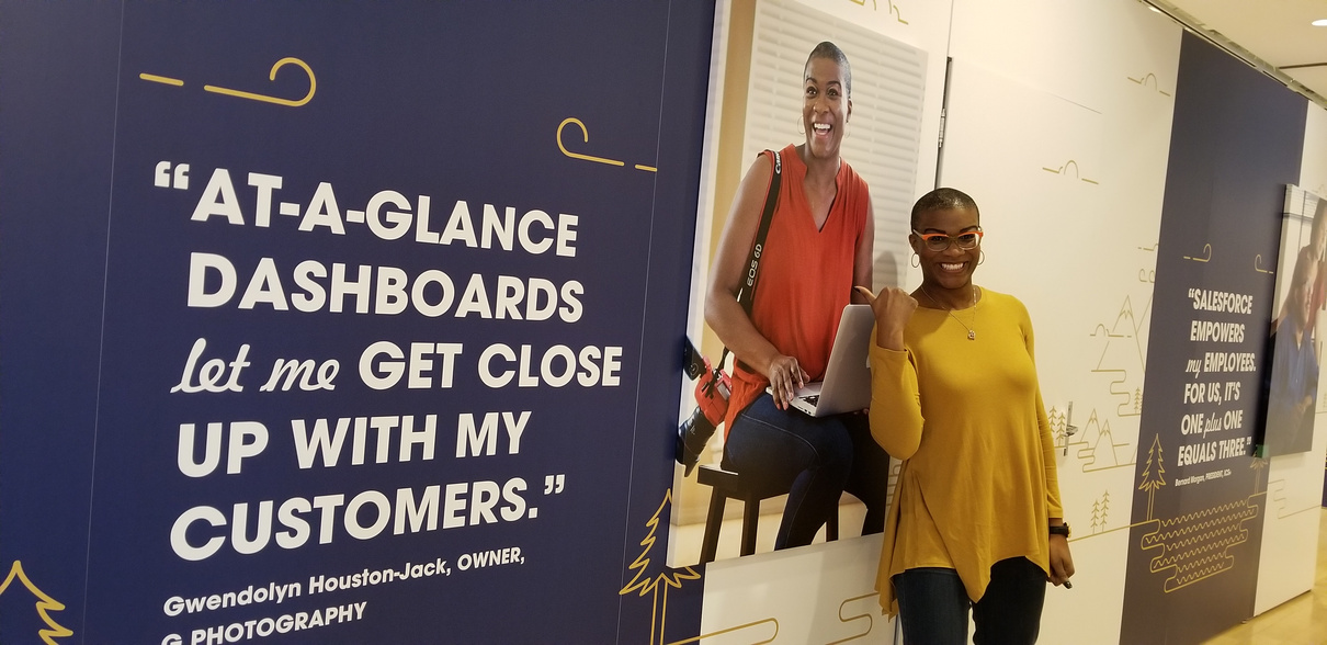 Gwendolyn R. Houston-Jack is a black woman photographer, creative, career coach and podcaster. She is pointing at her Salesforce advertisement at Salesforce Dreamforce 2019.